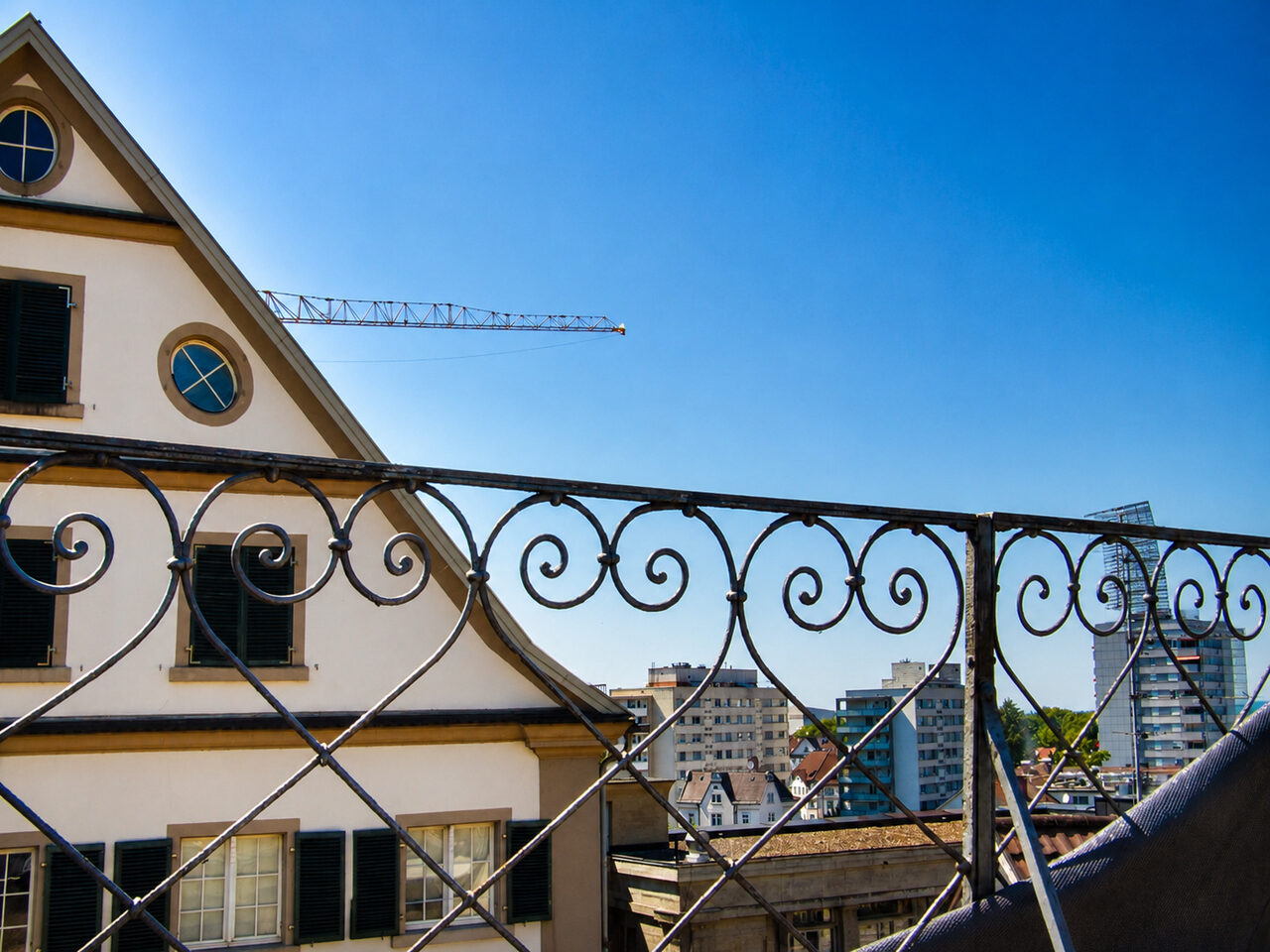 Blick vom Dach über ein schmiedeeisernes Geländer auf die Dächer von Bregenz, am Himmel ein Baukran
